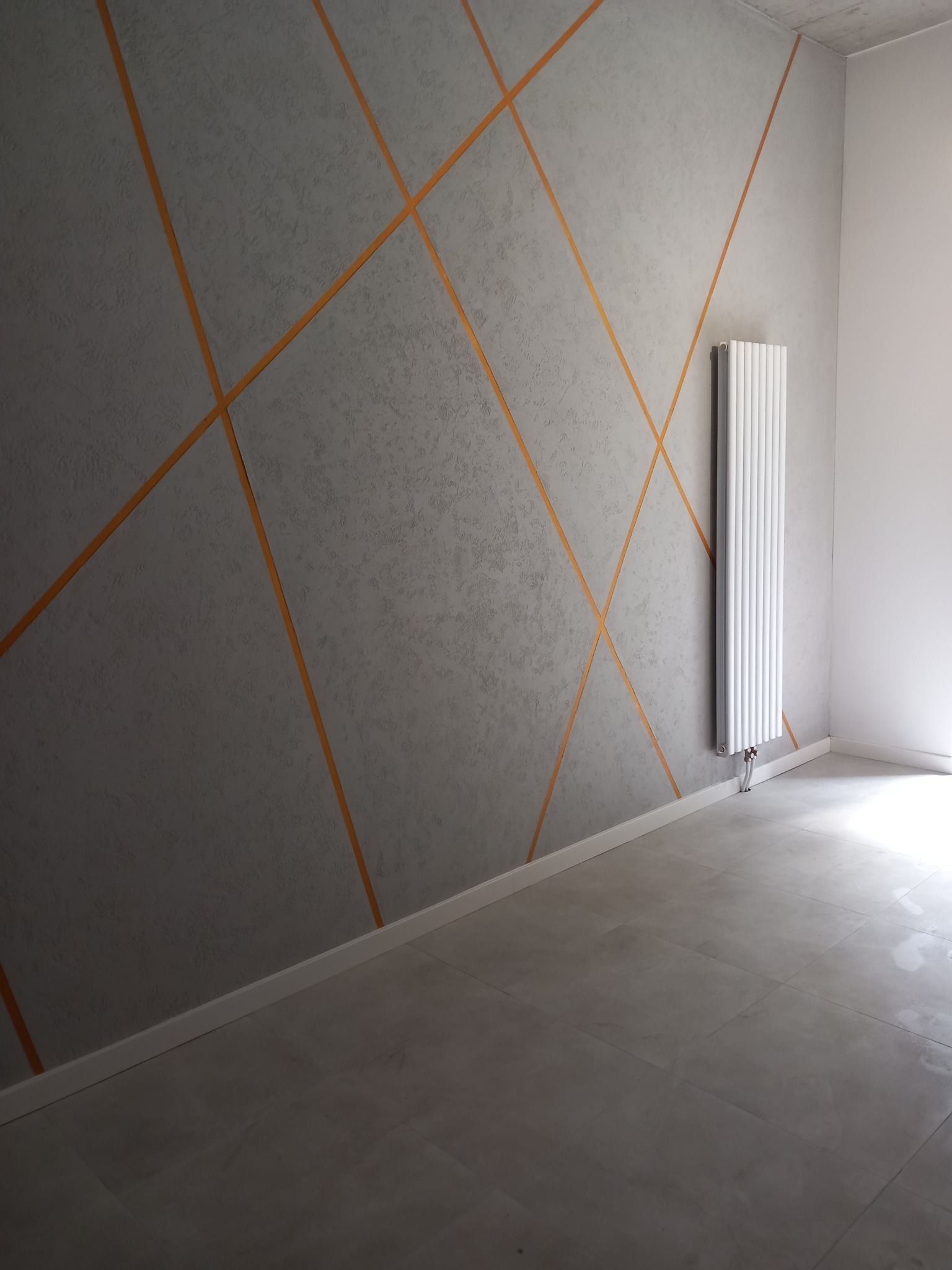 Szara ściana z dekoracyjnymi, pomarańczowymi liniami, grzejnikiem i jasną podłogą w pustym pokoju.