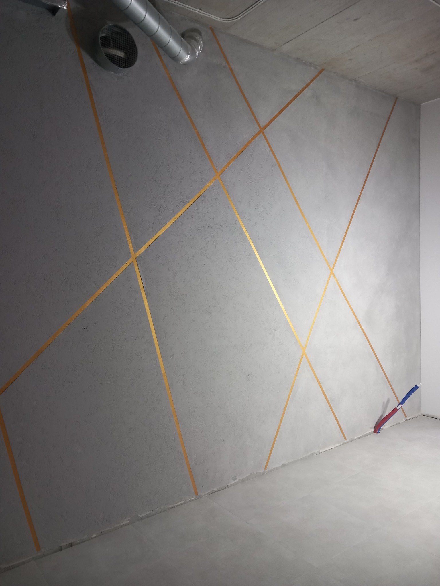 Szara ściana z dekoracyjnymi, złotymi paskami tworzącymi geometryczny wzór, widoczne instalacje wentylacyjne pod sufitem oraz rury przy podłodze.