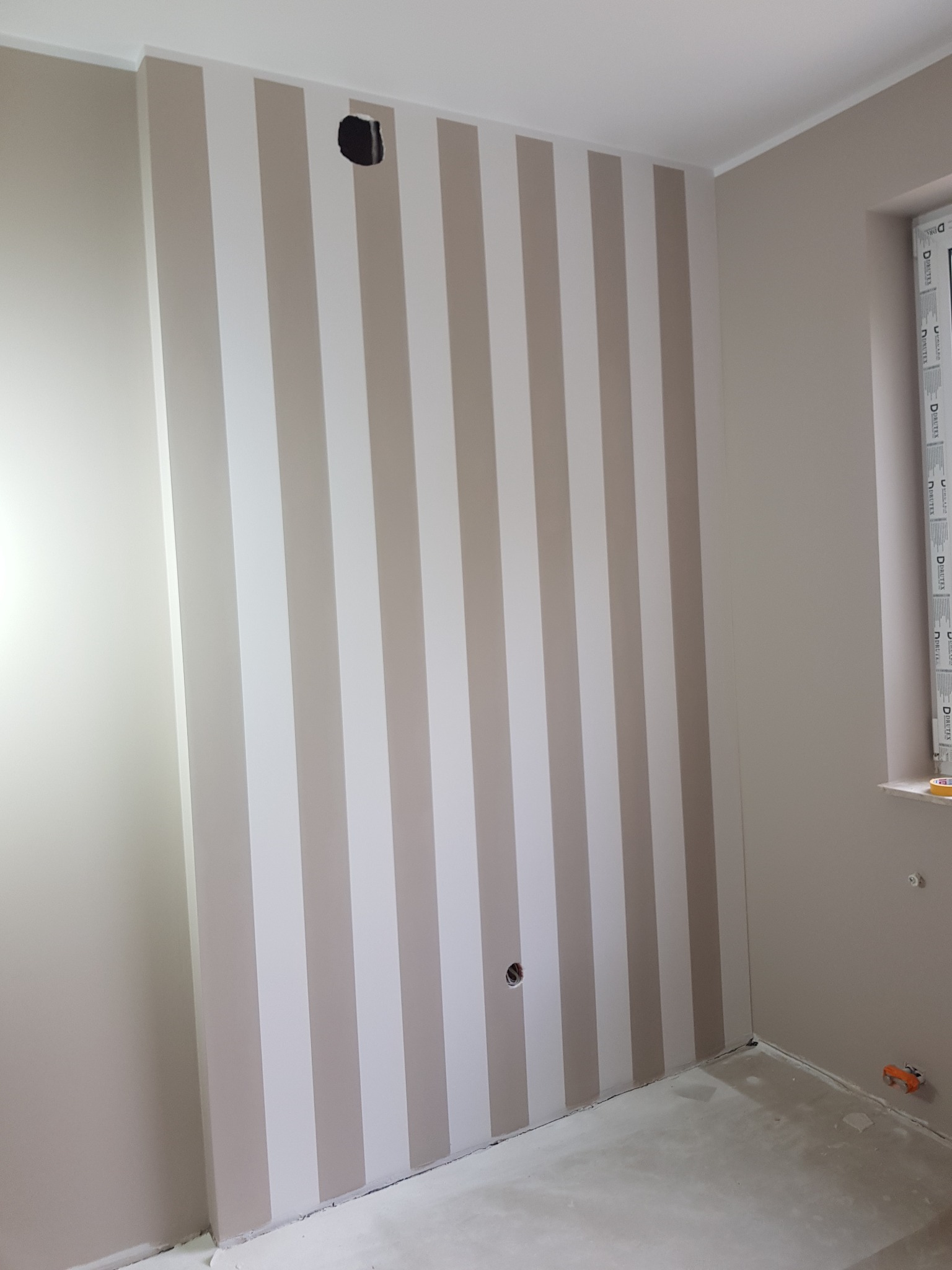 Ściana w pokoju pomalowana w pionowe pasy w odcieniach beżu i bieli, widoczne gniazdka elektryczne i surowa podłoga przed wykończeniem.