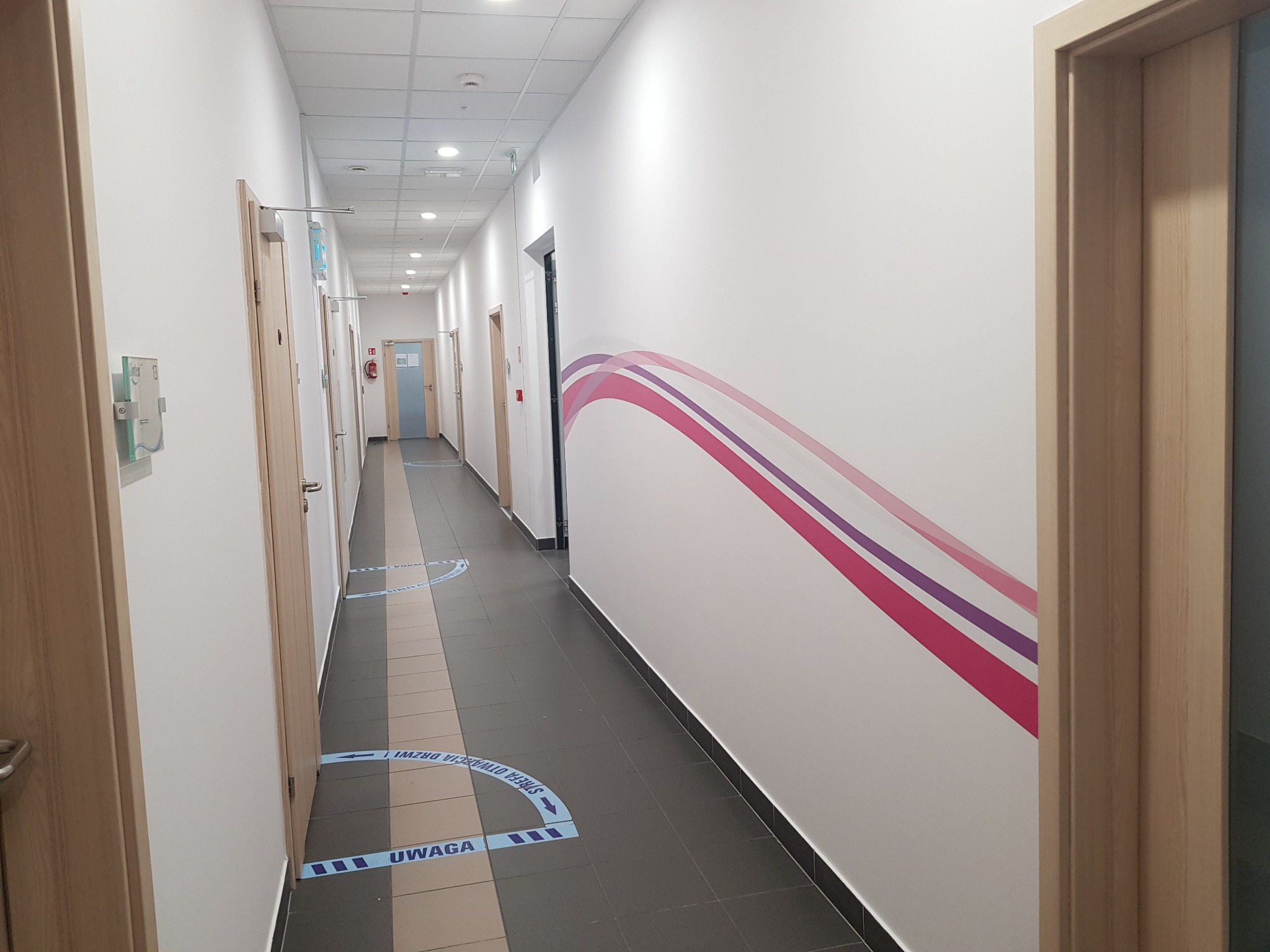 Długi korytarz biurowy z białymi ścianami, szarą podłogą z elementami beżu, drewnianymi drzwiami i dekoracyjnym motywem graficznym w odcieniach różu i fioletu na ścianie. Na podłodze umieszczone są...