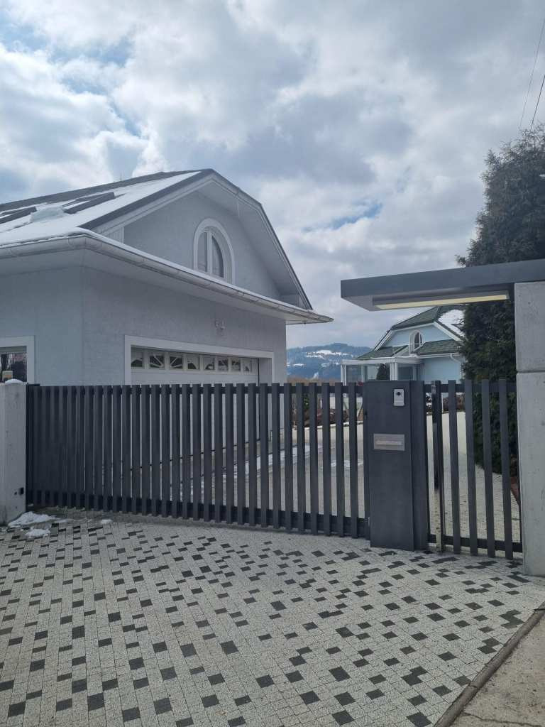 Nowoczesna, szara brama wjazdowa z pionowych profili z furtką, zamontowana na posesji z kostką brukową, w tle dom z białymi elementami i zaśnieżony dach.