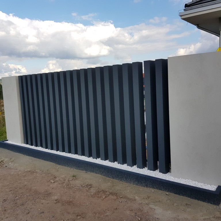 Nowoczesne ogrodzenie panelowe z pionowych, grafitowych profili, osadzone między dwoma betonowymi słupkami, z drobnym białym grysem u podstawy.