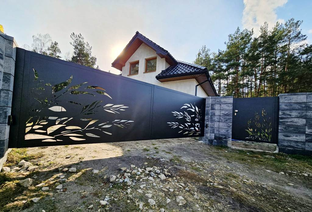 Nowoczesna, przesuwna brama metalowa w kolorze czarnym z laserowo wycinanym motywem roślinnym, osadzona na kamiennych słupkach, z widocznym domem w tle i nierównym podłożem.