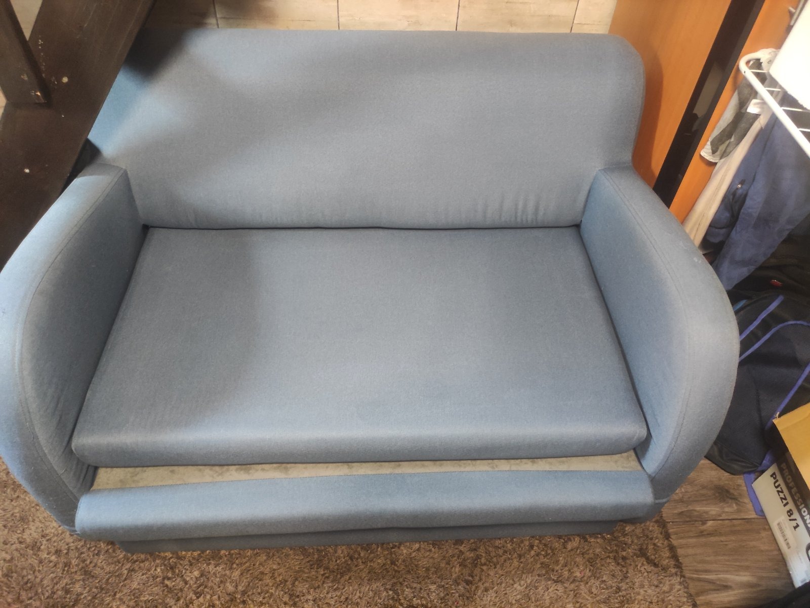 Niebieska, dwuosobowa sofa z zaokrąglonymi podłokietnikami, ustawiona na brązowym dywanie shaggy.