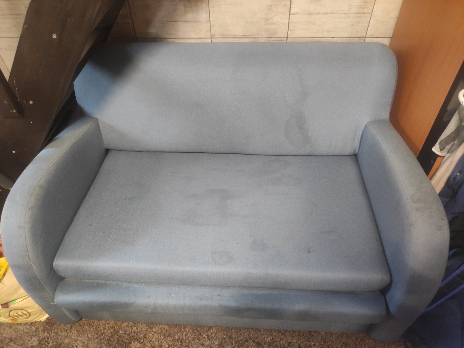 Szara, dwuosobowa kanapa z widocznymi plamami na tapicerce, ustawiona na brązowym dywanie, z fragmentem drewnianej konstrukcji w tle.