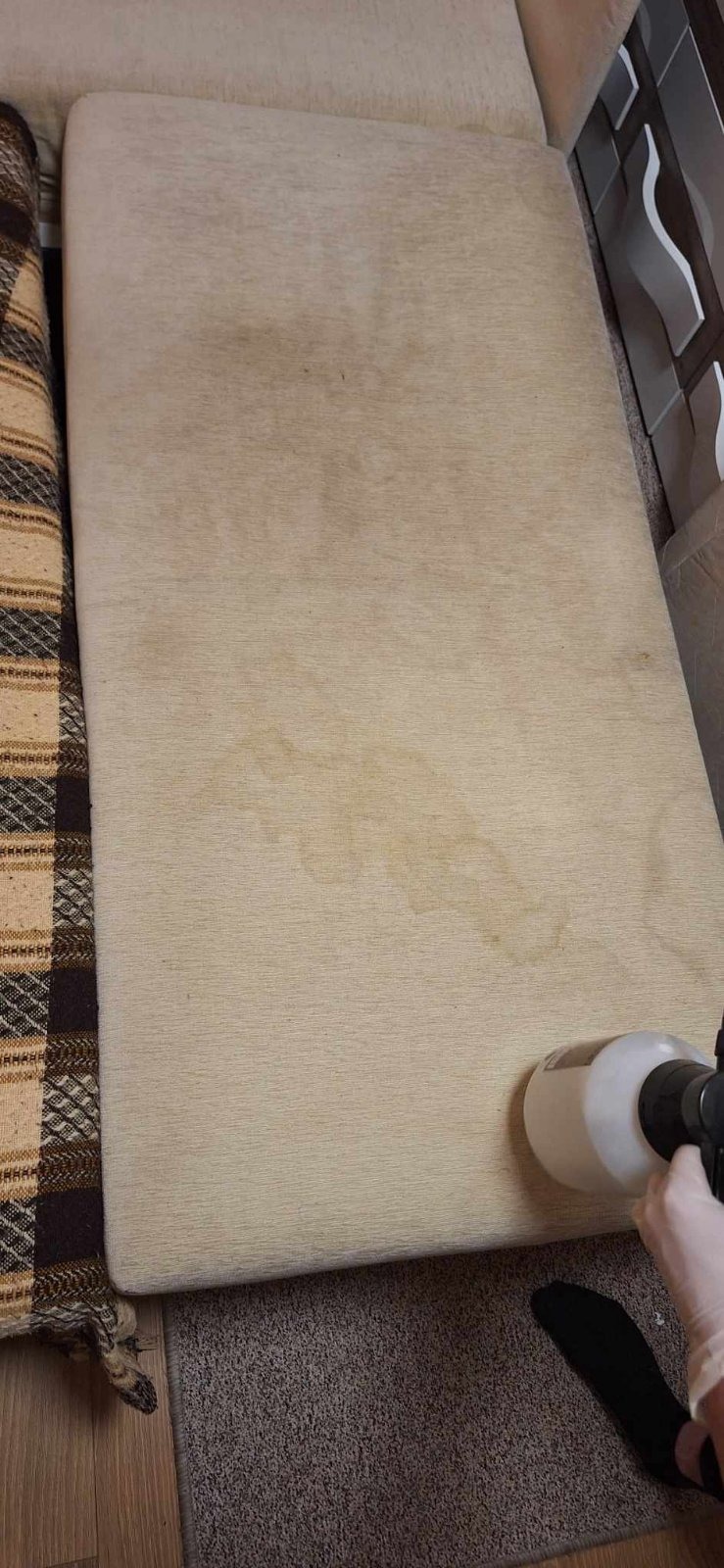 Czyszczenie beżowej kanapy z widocznymi plamami za pomocą spryskiwacza, ręka w lateksowej rękawicy, fragment wzorzystej narzuty w tle.