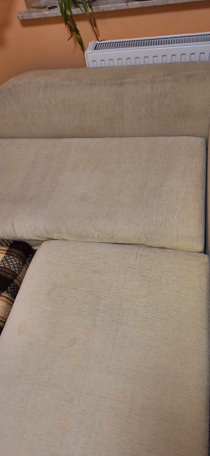 Fragment beżowej kanapy z widocznymi plamami na siedzisku i oparciu, obok leży brązowo-beżowy pled w kratę, w tle grzejnik