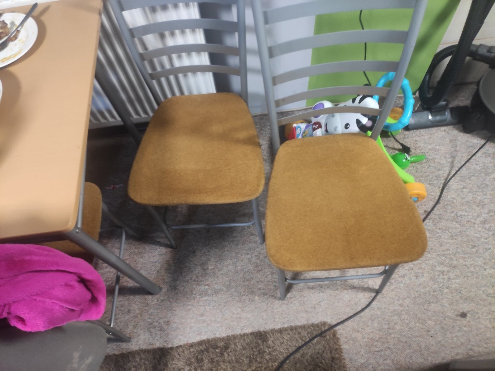 Dwa krzesła tapicerowane tkaniną w odcieniach brązu, z szarymi metalowymi ramami, stoją na dywanie. W tle widoczne zabawki dziecięce i odkurzacz.