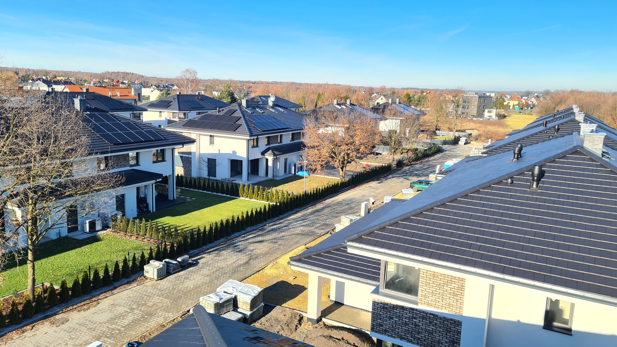 Nowoczesne domy jednorodzinne z panelami słonecznymi na dachach, widok z góry na osiedle w słoneczny dzień, widoczne kostki brukowe i przygotowany teren.