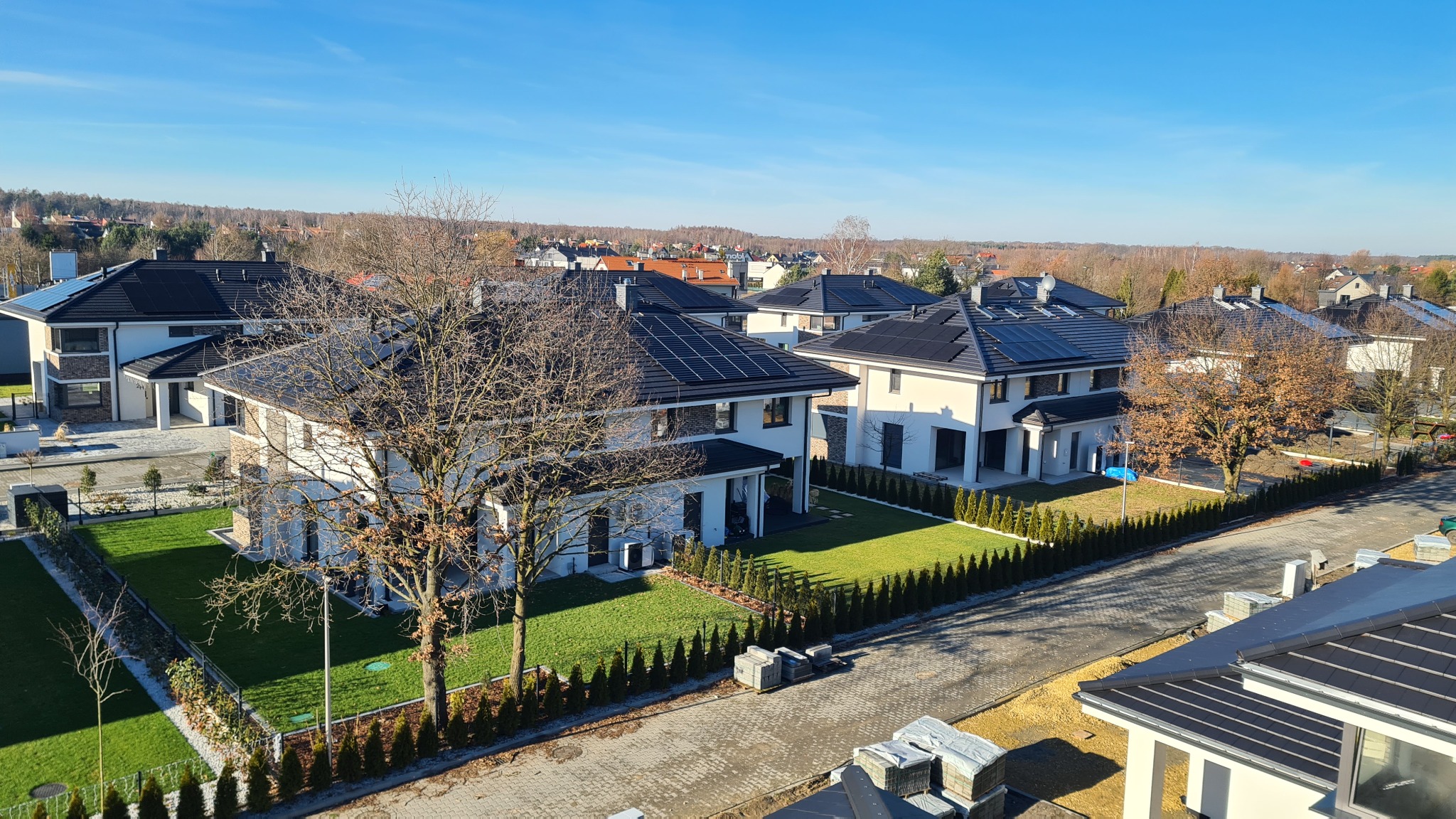 Nowoczesne domy jednorodzinne z białymi elewacjami i panelami słonecznymi na dachach, widok z góry na osiedle w słoneczny dzień, z widocznymi drzewami bez liści i kostką brukową na ulicy.