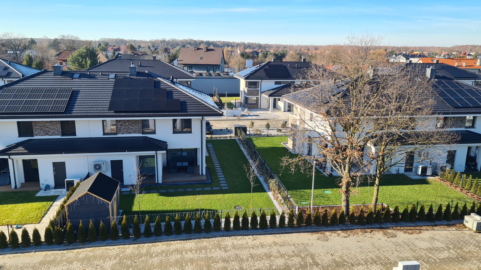 Nowoczesne, białe domy jednorodzinne z czarnymi dachami i panelami słonecznymi w słoneczny dzień, widok z góry na zadbane trawniki i niskie żywopłoty.