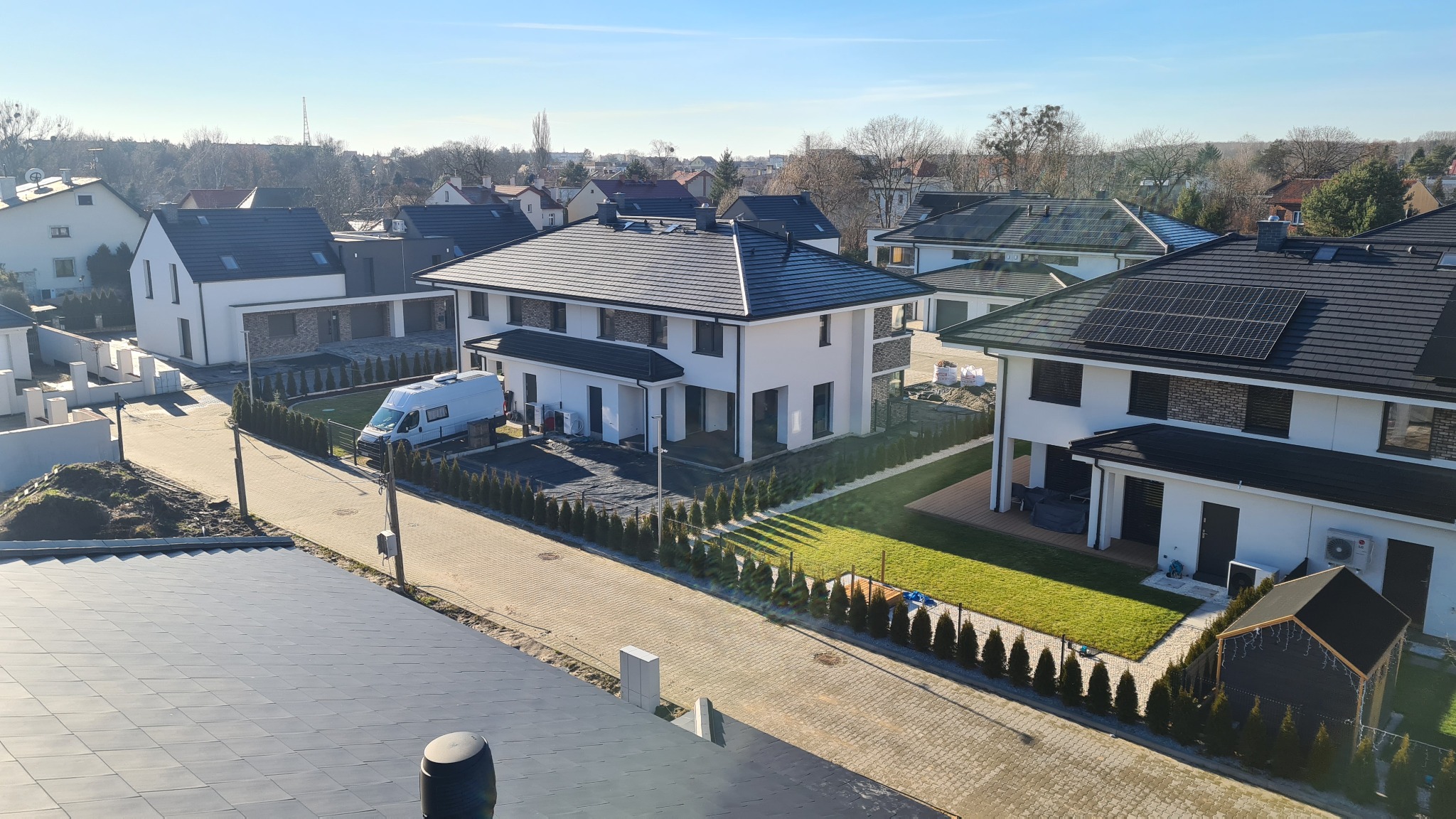 Panorama osiedla domów jednorodzinnych z białymi elewacjami, czarnymi dachami z panelami solarnymi i starannie utrzymanymi trawnikami, widziana z góry w słoneczny dzień, z zaparkowanym białym vanem...