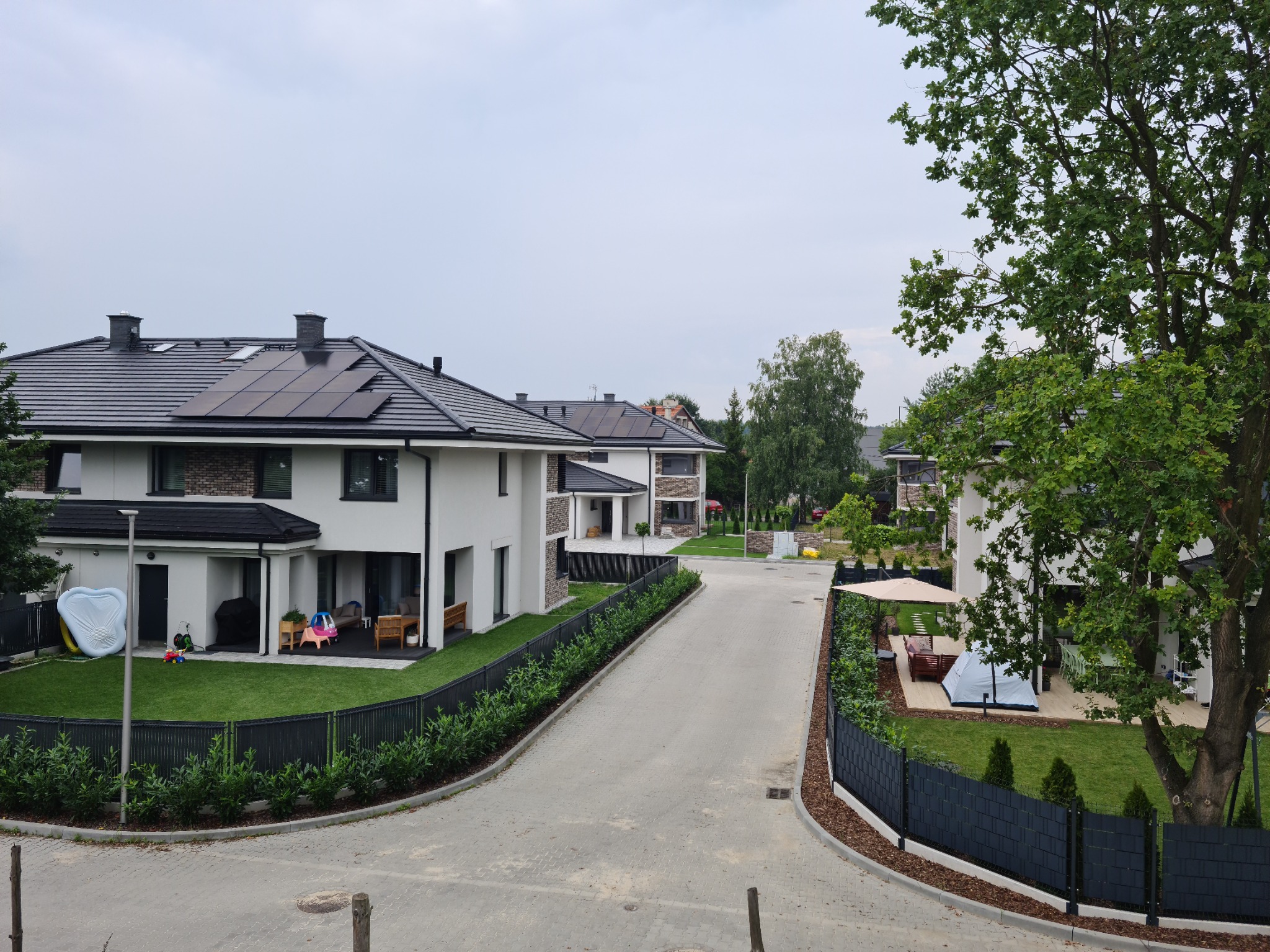 Nowoczesne domy jednorodzinne z białymi elewacjami, czarnymi dachami z panelami słonecznymi i zadbanymi trawnikami, widok z góry na osiedle.