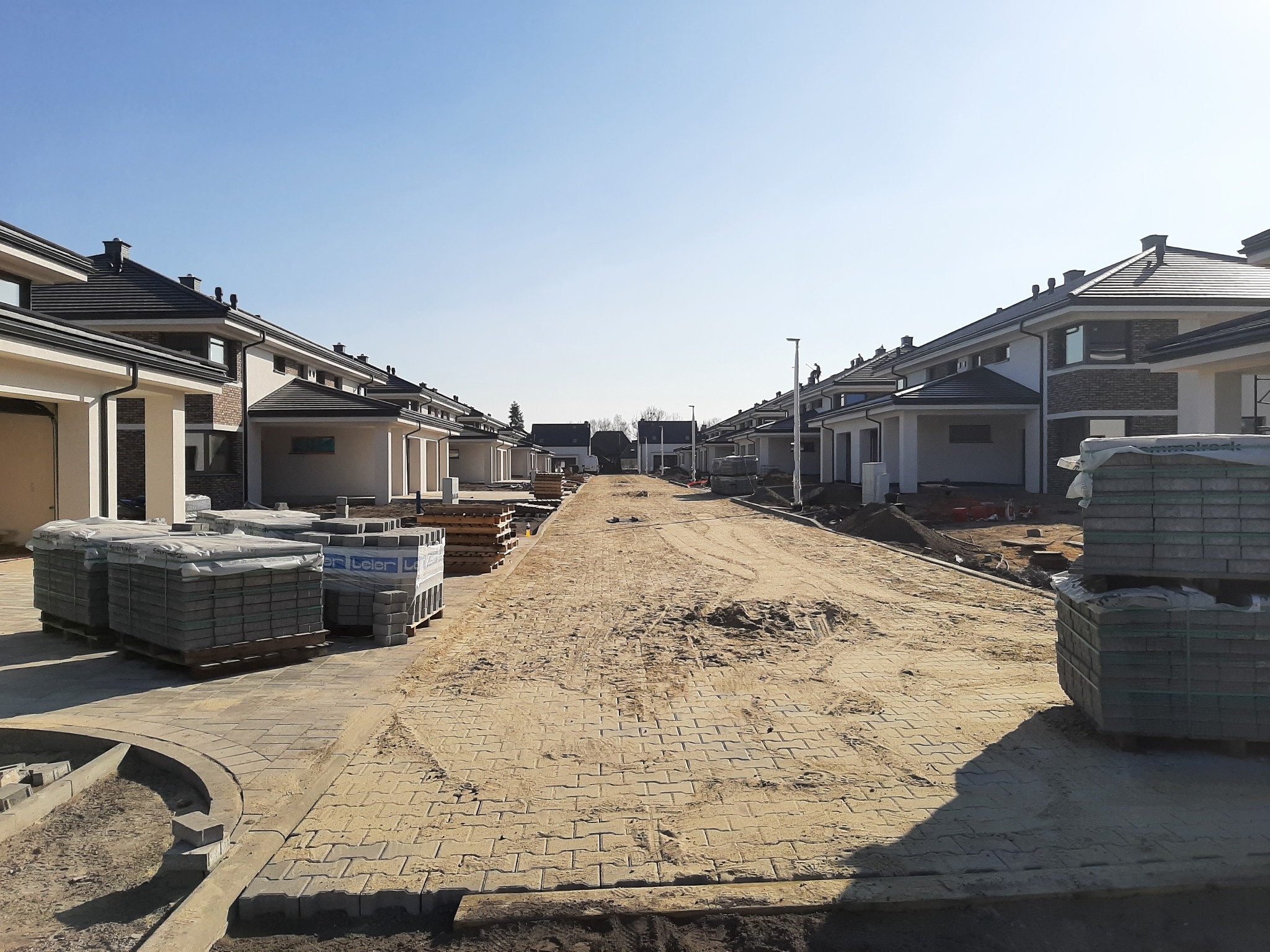 Nowe osiedle domów jednorodzinnych w trakcie budowy, widoczne palety z kostką brukową i częściowo ułożony chodnik.
