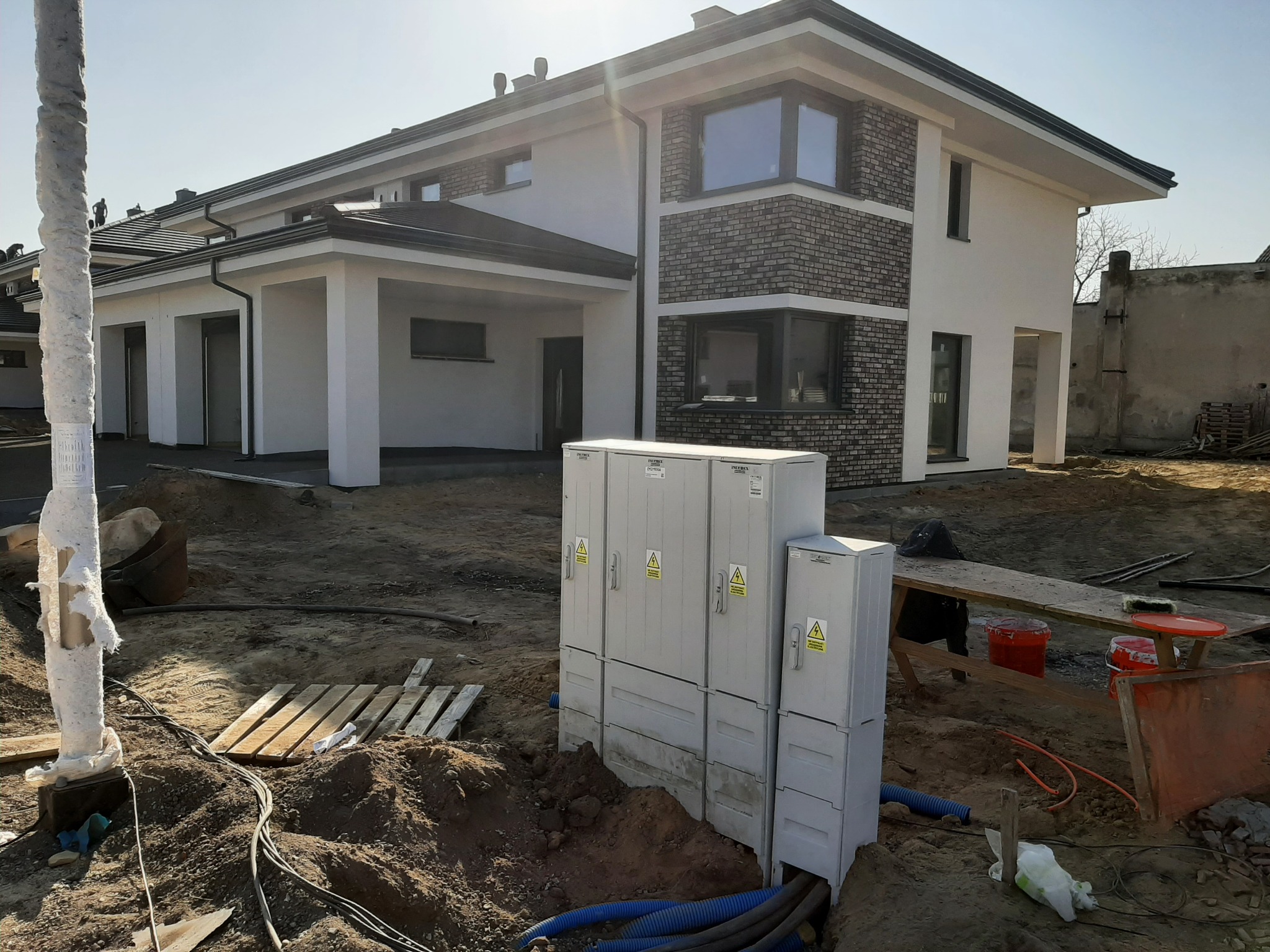 Nowoczesny dom jednorodzinny z elementami cegły klinkierowej na elewacji, widoczne skrzynki elektryczne i teren w trakcie prac wykończeniowych.