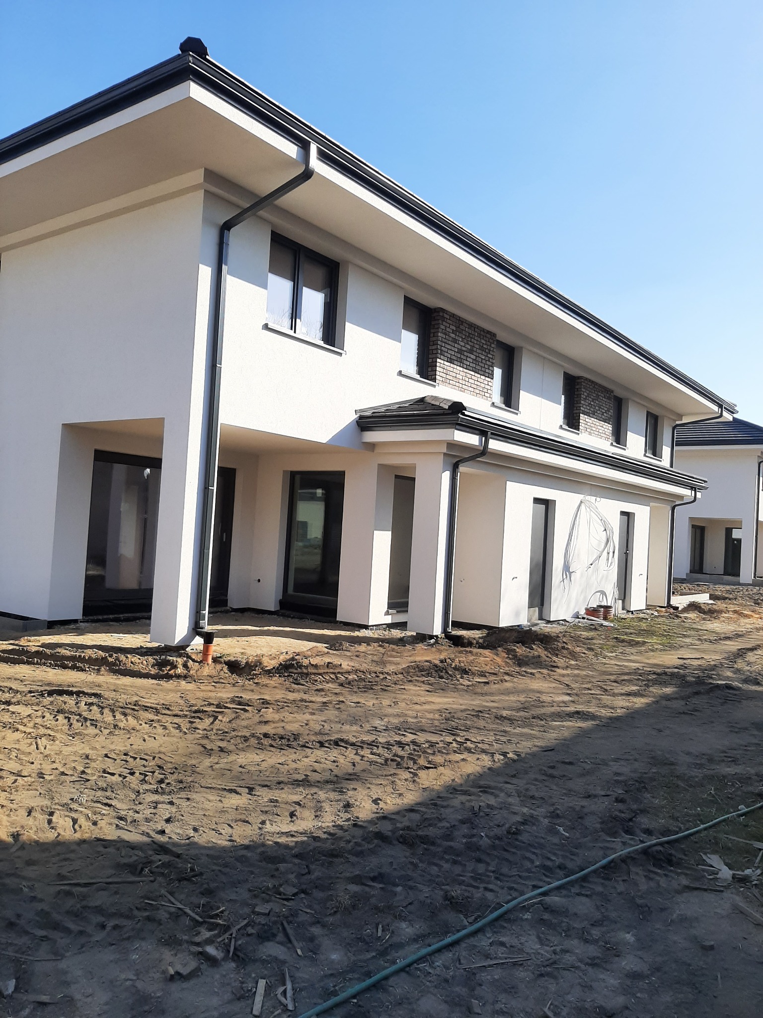 Nowoczesny, dwukondygnacyjny dom w zabudowie szeregowej z białą elewacją, czarnymi oknami i orynnowaniem, teren wokół domu w trakcie prac ziemnych, widoczne ślady opon i rury.