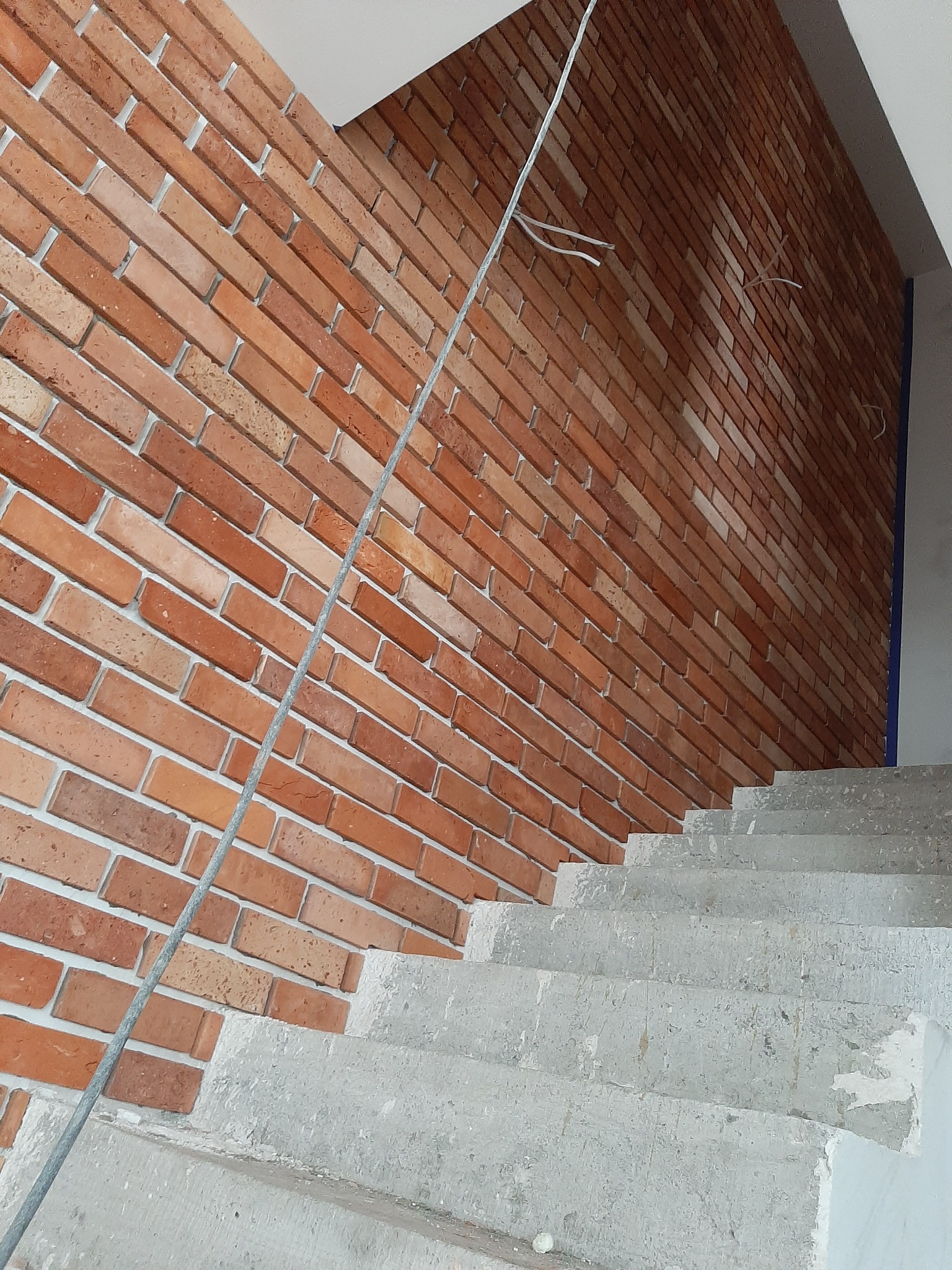 Ściana wykończona czerwoną cegłą klinkierową z widocznymi spoinami, obok surowe betonowe schody w trakcie budowy, z wiszącym kablem elektrycznym.