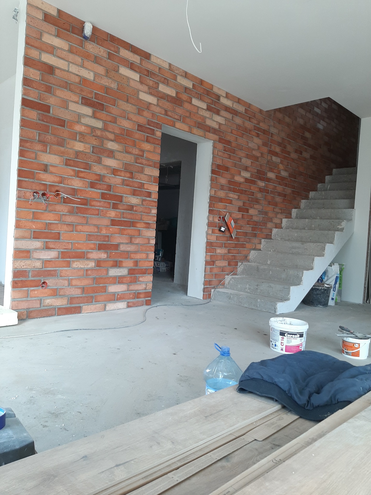 Wnętrze w trakcie wykańczania: ściana wyłożona cegłą klinkierową, surowe betonowe schody, widoczne puszki z zaprawą i narzędzia budowlane, instalacja elektryczna bez osprzętu.