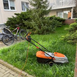 NOVA CLEAN VITALII TSYTSYLIN - Pomarańczowa kosiarka elektryczna na trawniku przed blokiem w Warszawie. Obok zaparkowane rowery, w tle iglaste drzewo i balkon.