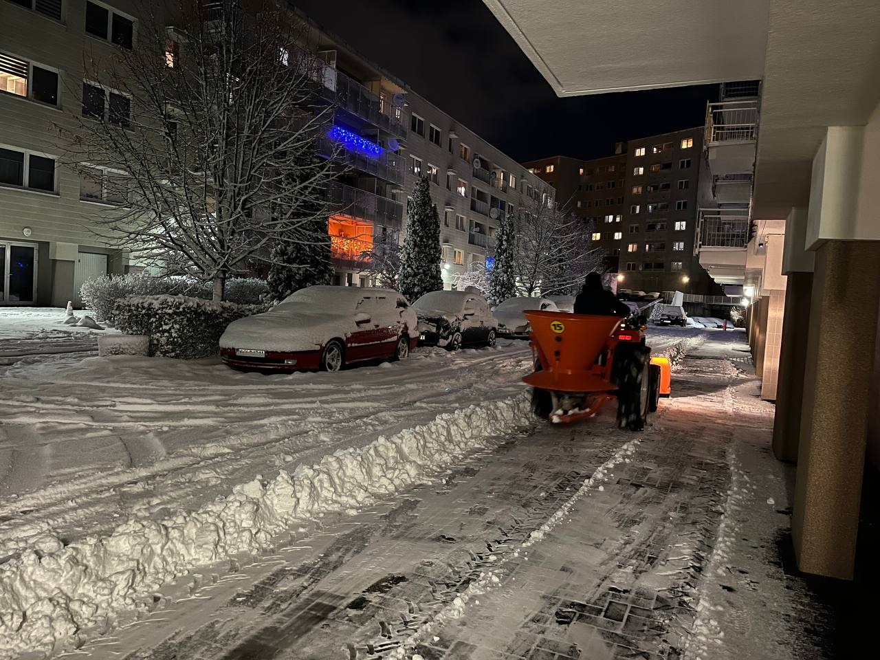 Pomarańczowy traktor odśnieża chodnik w zimowy wieczór. Śnieg pokrywa samochody i drzewa na osiedlu mieszkaniowym. Widoczne ślady opon na śniegu.
