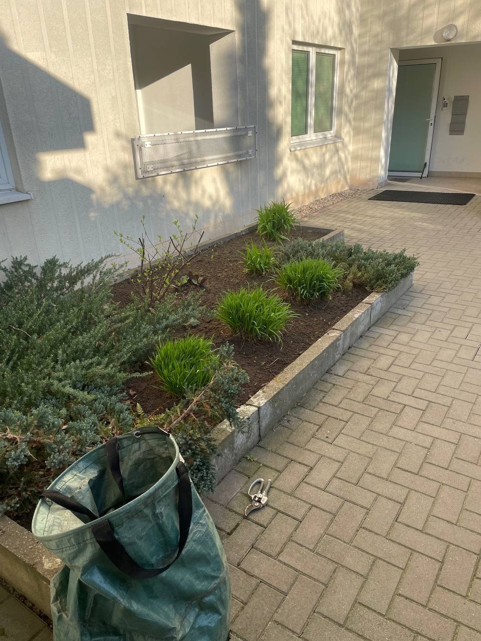 Kwitnąca rabata kwiatowa przy budynku, z torbą ogrodniczą i sekatorem na kostce brukowej. Świeżo posadzone rośliny w wiosennym słońcu.