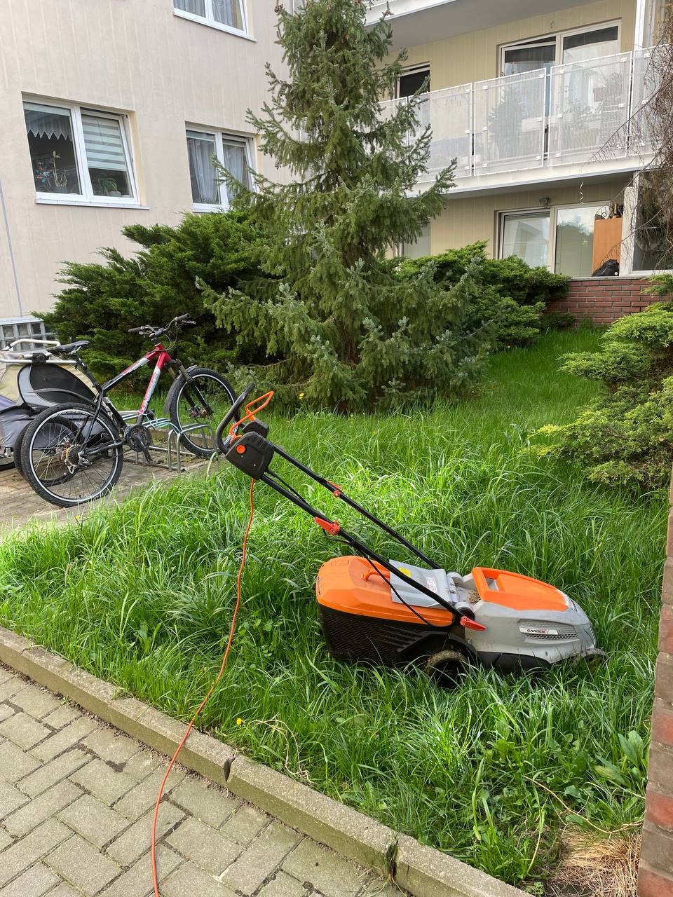 Pomarańczowa kosiarka elektryczna na trawniku przed blokiem w Warszawie. Obok zaparkowane rowery, w tle iglaste drzewo i balkon.
