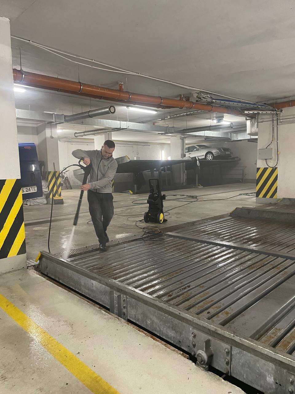 Mężczyzna czyści myjką ciśnieniową metalową platformę w garażu podziemnym. W tle zaparkowane samochody i instalacje pod sufitem.