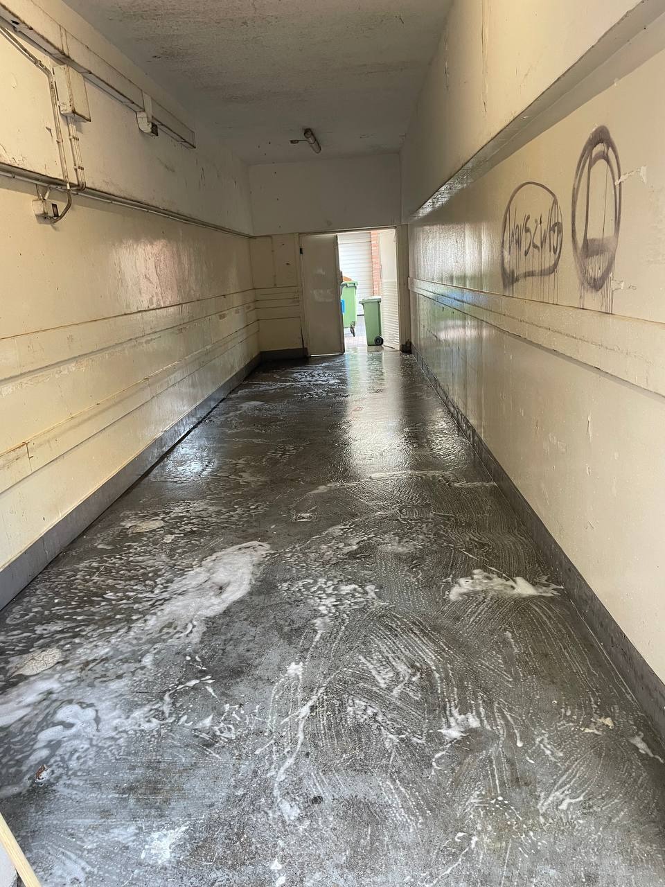 Korytarz w trakcie mycia podłogi, widoczne ślady detergentu i graffiti na ścianie. W tle zielone kosze na śmieci.