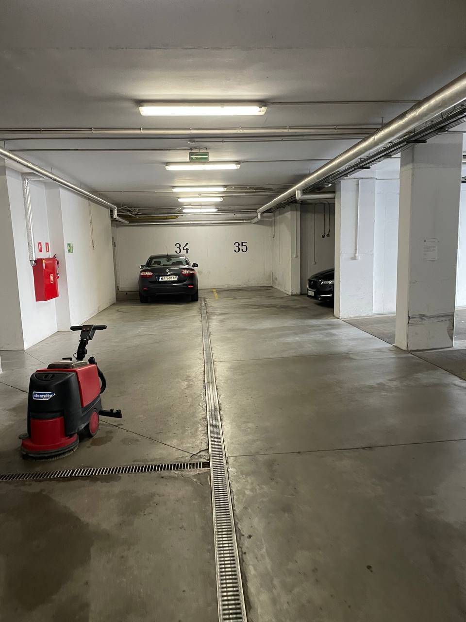 Czerwono-czarna maszyna czyszcząca Cleanfix na betonowej posadzce parkingu podziemnego z numerami miejsc 34 i 35 w tle. Widoczne odpływy liniowe.