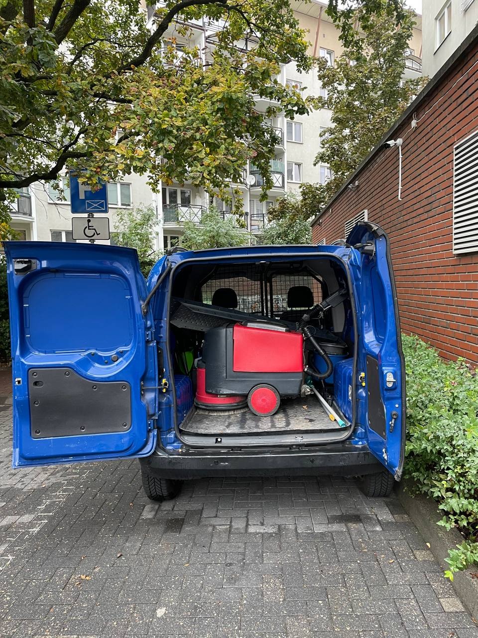 Otwarty, niebieski van z czerwoną maszyną do czyszczenia podłóg w środku, zaparkowany na zewnątrz budynku z cegły i zielenią. Sprzęt do sprzątania.