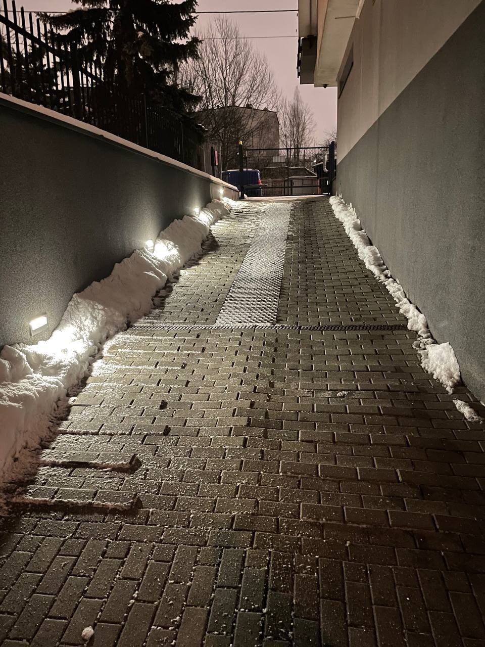 Zimowy wjazd do budynku z kostki brukowej, oświetlony lampami. Śnieg zalega po bokach, a w tle widoczny budynek i drzewa.