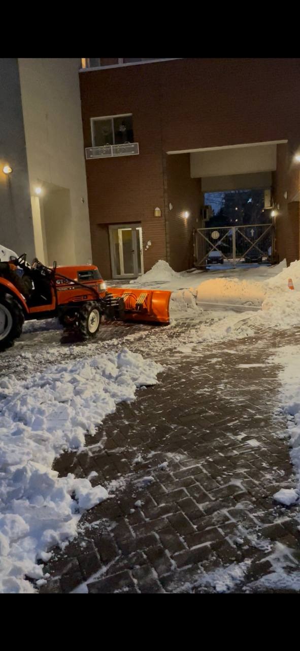 Pomarańczowy traktor z pługiem odśnieża kostkę brukową przed budynkiem. Widoczne oświetlenie zewnętrzne i brama wjazdowa.