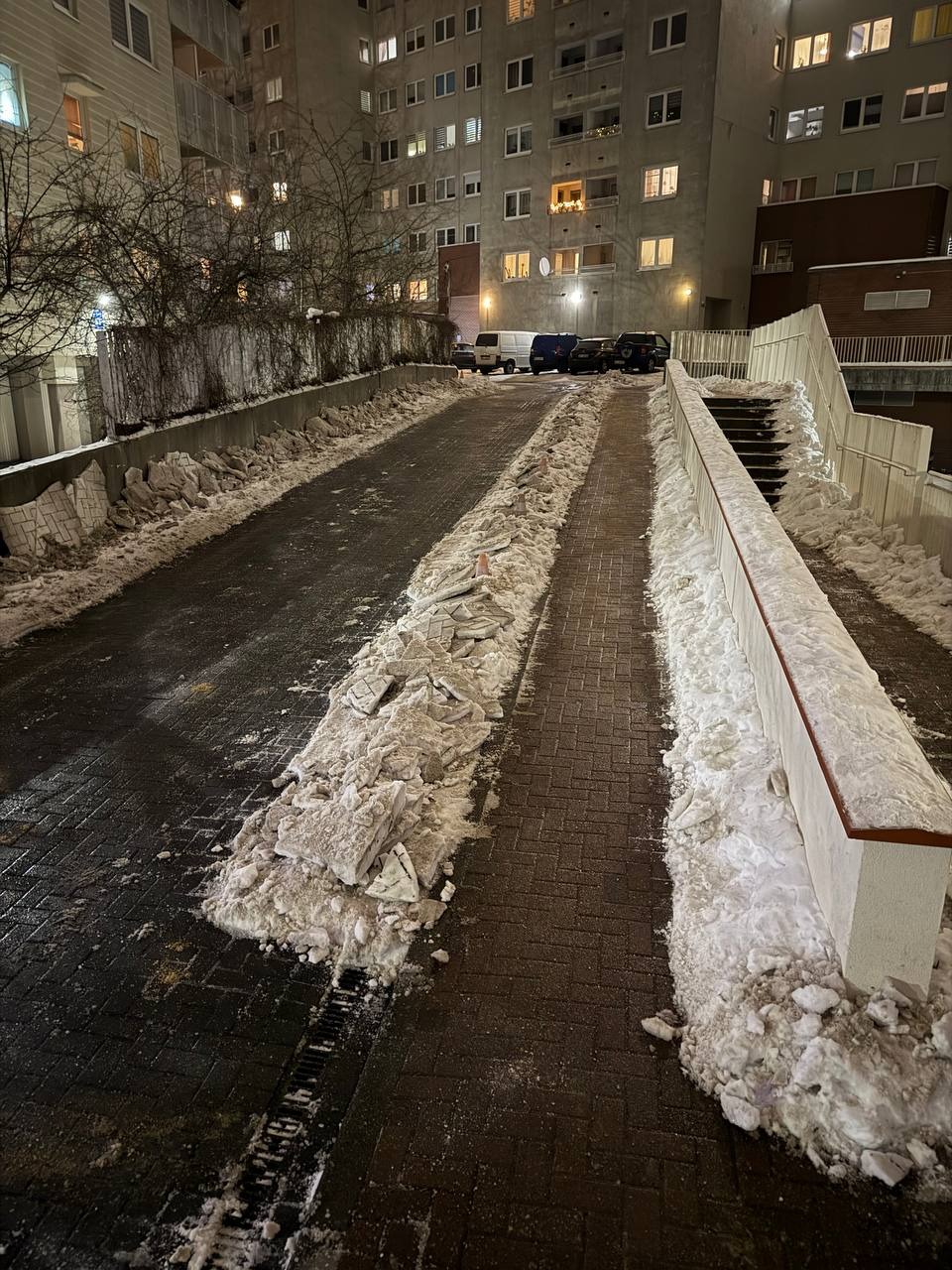 Zimowy krajobraz miejski: odśnieżona ścieżka z kostki brukowej prowadzi w kierunku bloku mieszkalnego, z zaspami śniegu po bokach i zaparkowanymi autami w tle.