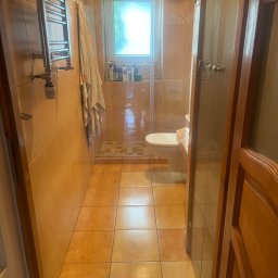 Łowca Kurzu - Umywalka i toaleta w łazience z beżowymi płytkami, widok zza uchylonych drewnianych drzwi. W tle kabina prysznicowa z przezroczystymi drzwiami i szklanymi półkami z kosmetykami.