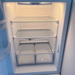 Łowca Kurzu - Wnętrze czystej, pustej lodówki z szklanymi półkami i przezroczystymi pojemnikami na warzywa, oświetlone wewnętrznym światłem.