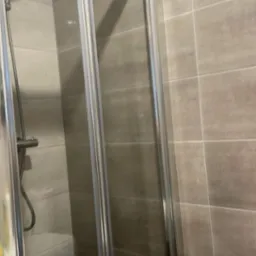 Szklana kabina prysznicowa z przesuwnymi drzwiami, widoczna słuchawka prysznicowa i płytki w odcieniach szarości, w tle pralka.