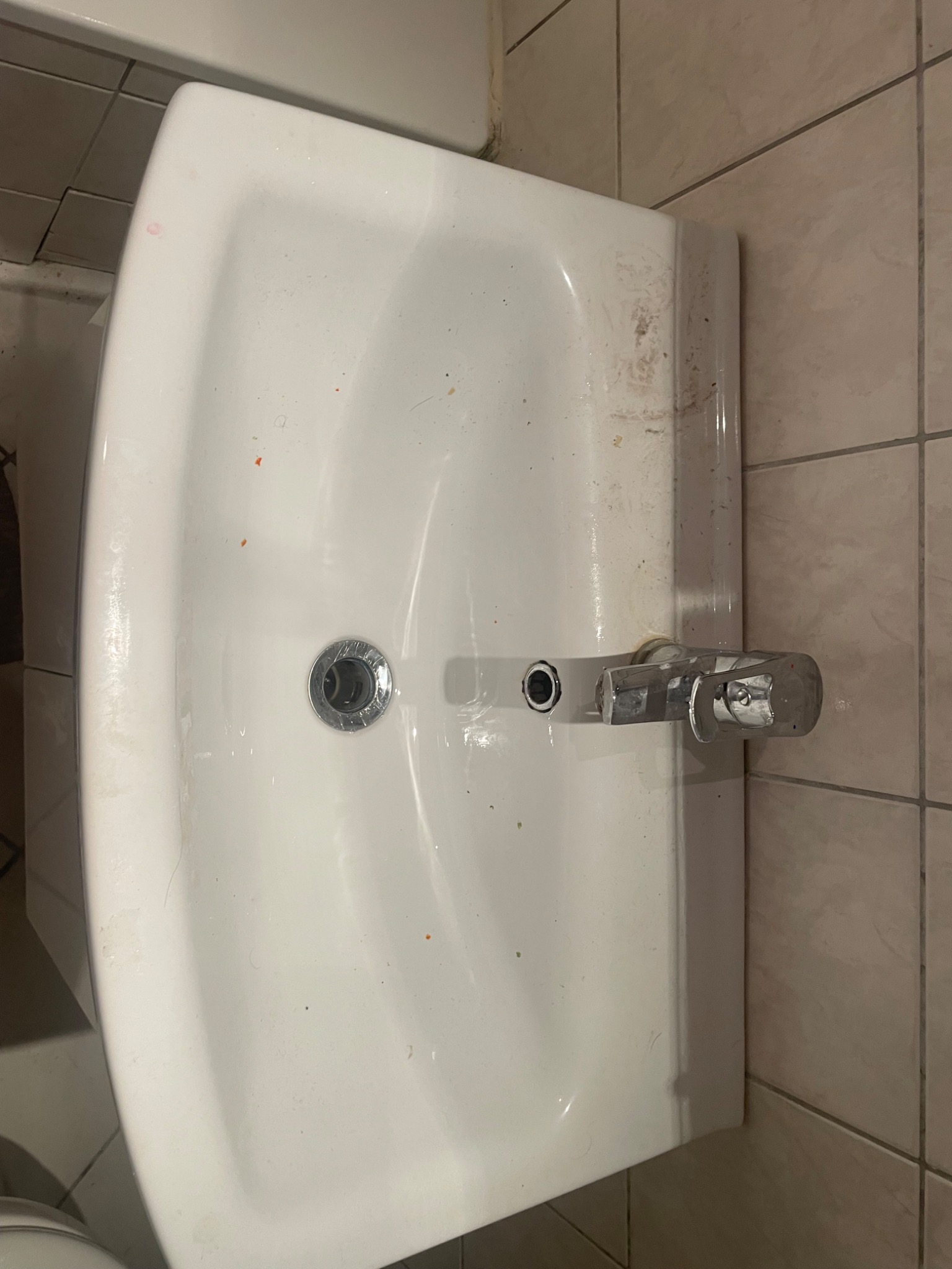 Brudna, biała umywalka z plamami i zabrudzeniami, widoczny kran i odpływ, płytki na ścianie w tle.