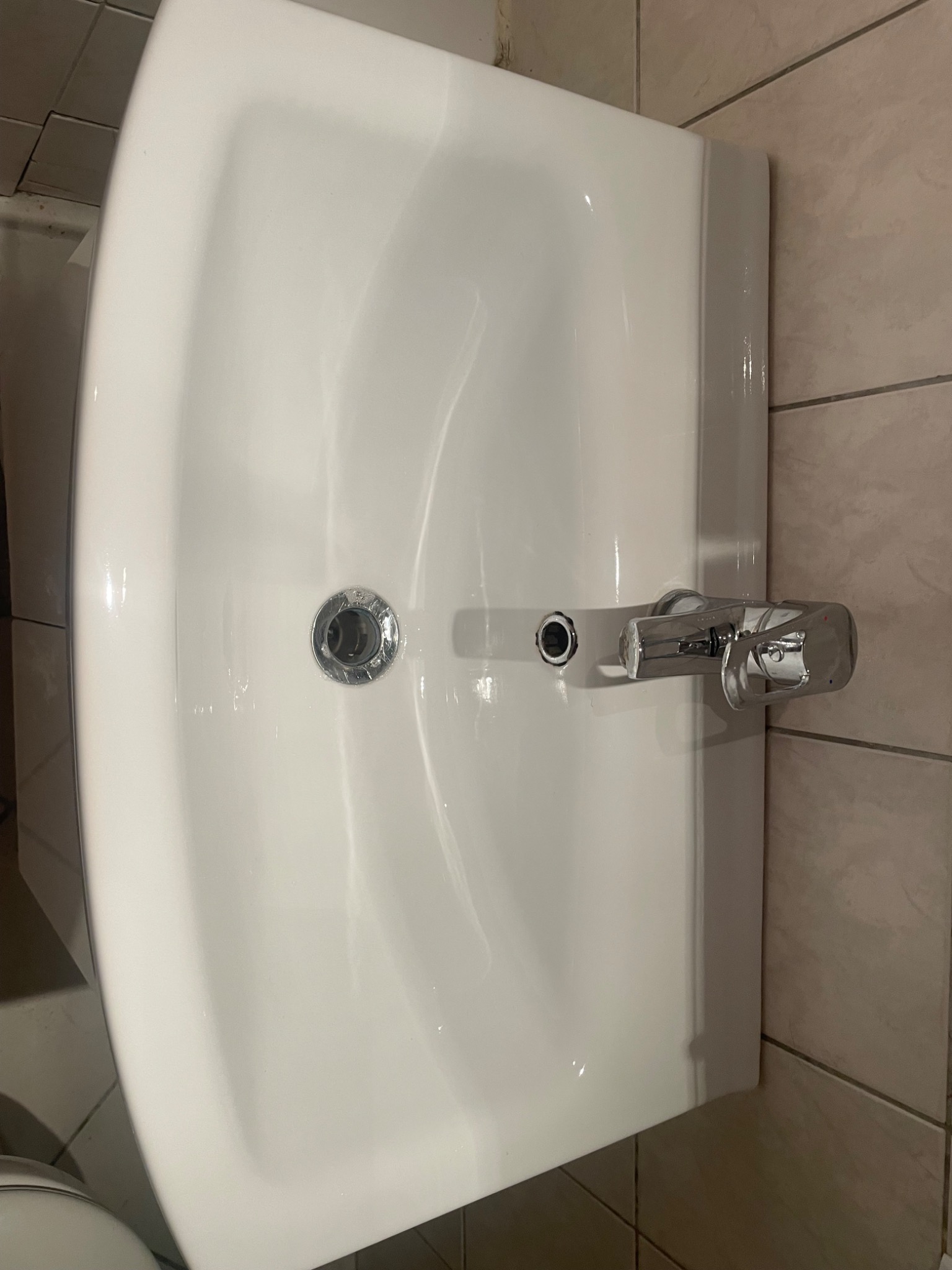 Biała umywalka z chromowaną baterią na tle beżowych płytek, widok z góry.