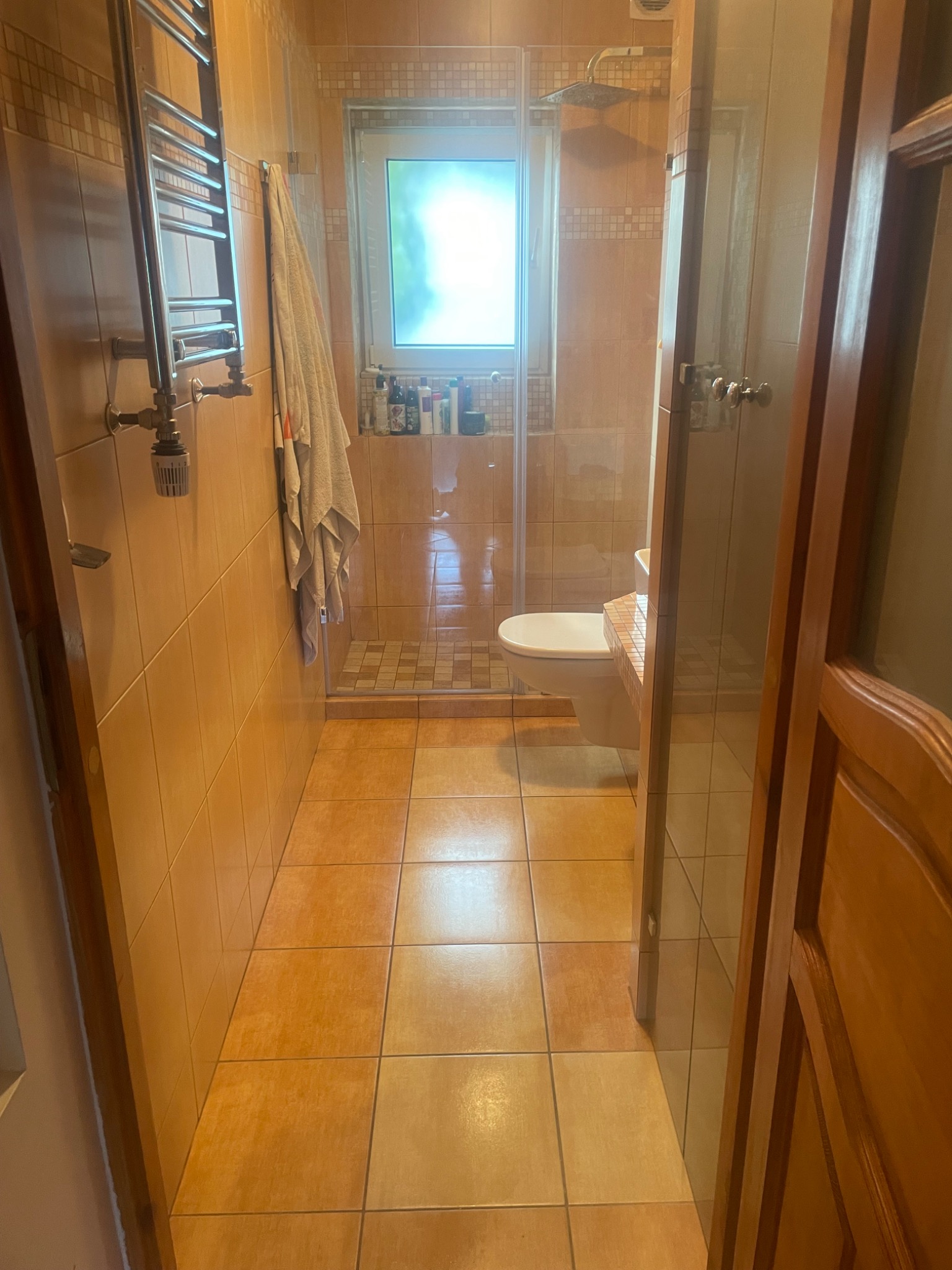 Umywalka i toaleta w łazience z beżowymi płytkami, widok zza uchylonych drewnianych drzwi. W tle kabina prysznicowa z przezroczystymi drzwiami i szklanymi półkami z kosmetykami.