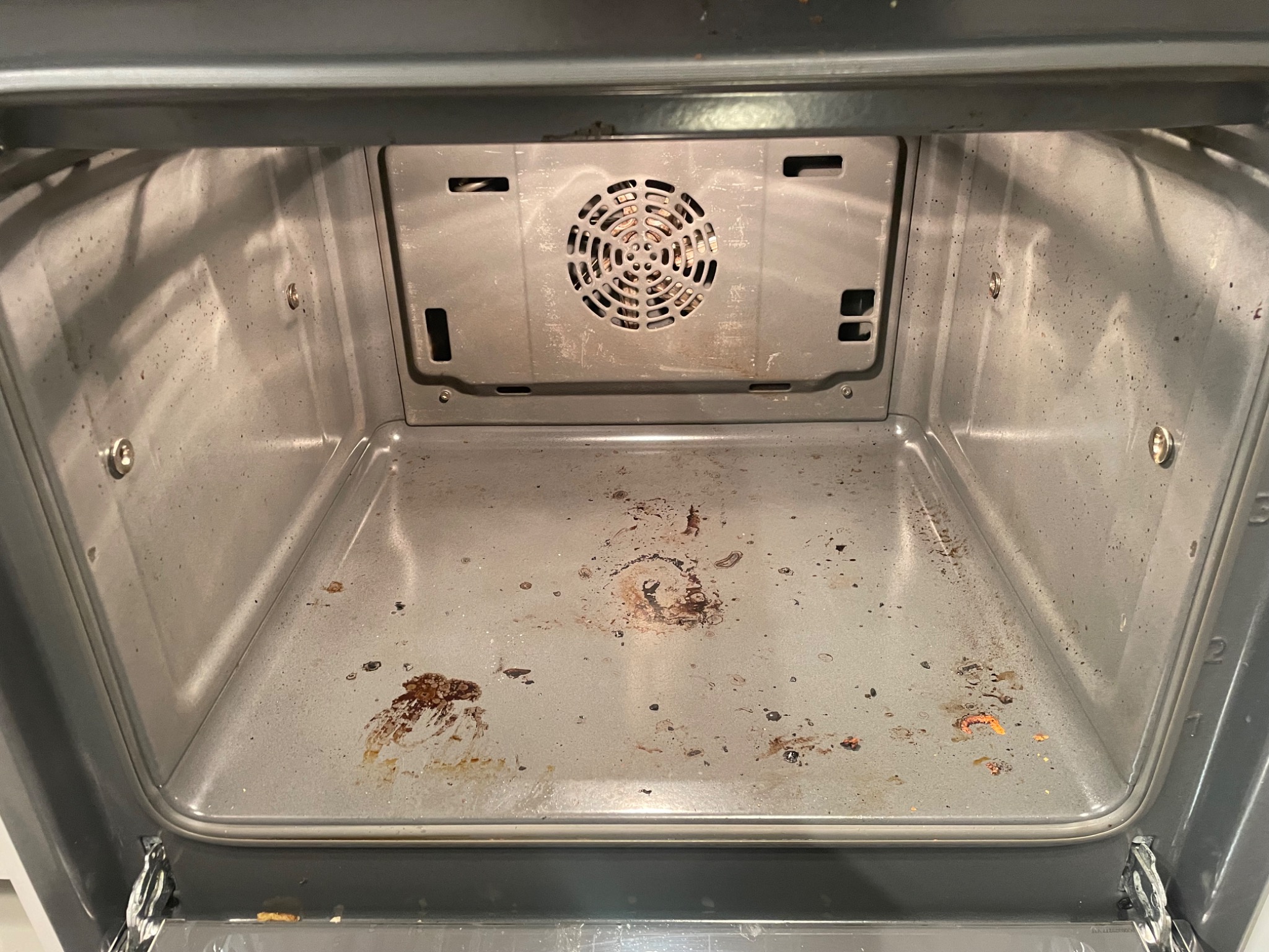 Wnętrze brudnego piekarnika z przypalonymi resztkami jedzenia na dnie i ściankach, widoczny wentylator z tyłu.