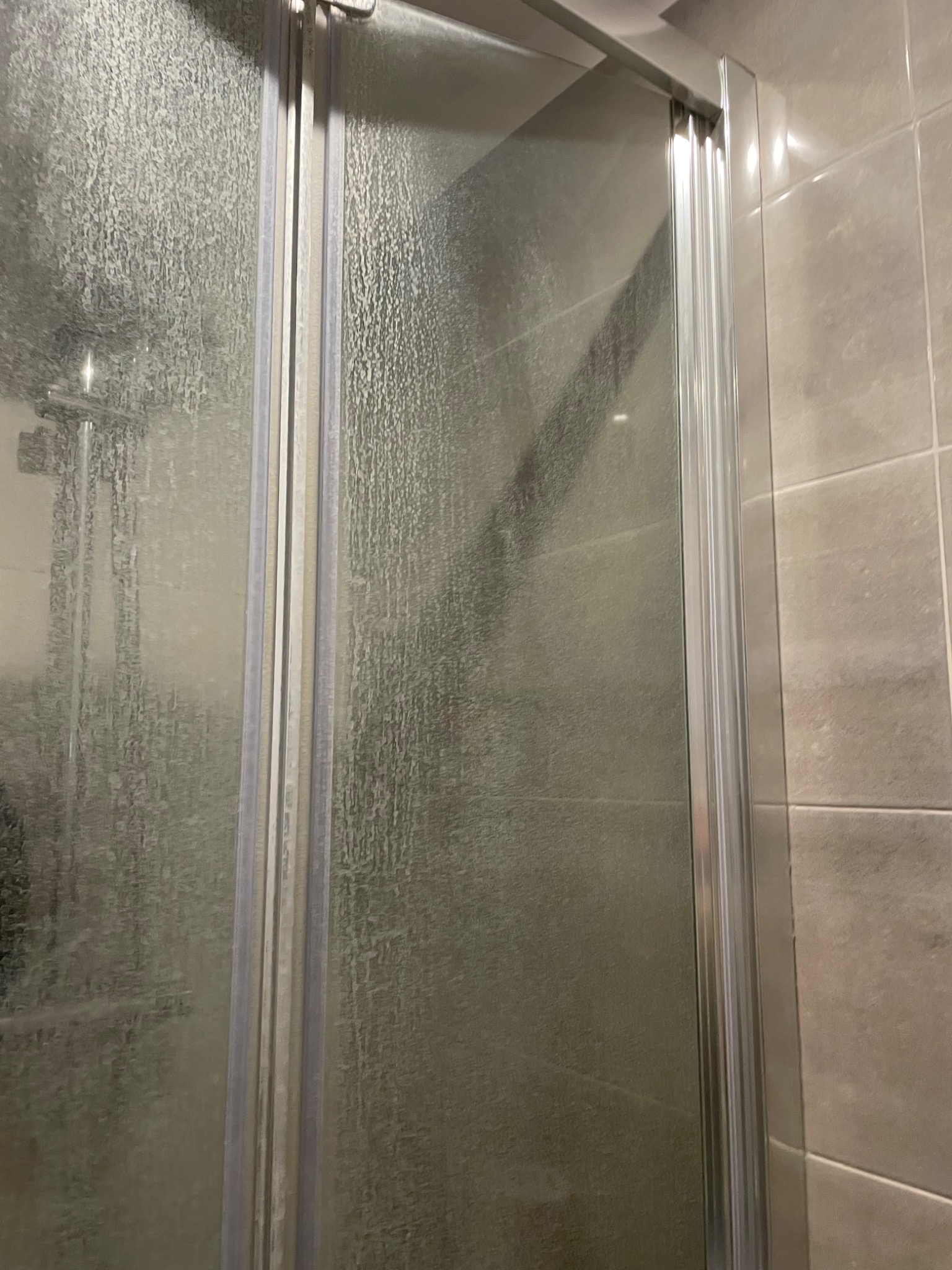 Zabrudzona kabina prysznicowa z widocznymi zaciekami i osadem z mydła na szklanych drzwiach, z fragmentem płytek w tle.