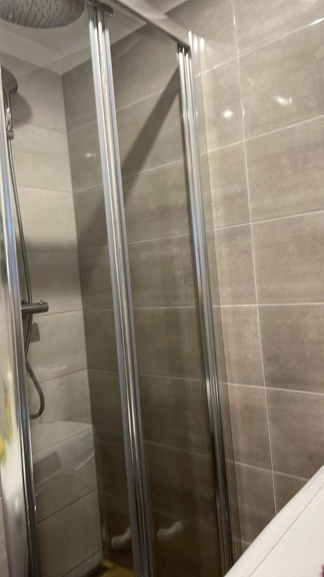 Szklana kabina prysznicowa z przesuwnymi drzwiami, widoczna słuchawka prysznicowa i płytki w odcieniach szarości, w tle pralka.