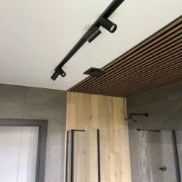 Nowoczesna łazienka z kabiną prysznicową typu walk-in, drewnianą okładziną ściany i sufitu oraz czarnym oświetleniem szynowym.