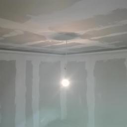 SUFITY - MP MARIUSZ PACZKOWSKI - Wnętrze w trakcie prac wykończeniowych: sufit i ściany pokryte wstępną warstwą szpachli, widoczna wisząca żarówka oświetlająca pomieszczenie. W tle widoczna listwa sufitowa.