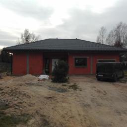 SUFITY - MP MARIUSZ PACZKOWSKI - Surowy budynek jednorodzinny z czerwoną elewacją w trakcie prac wykończeniowych, widoczny samochód dostawczy na podjeździe i materiały budowlane składowane obok.