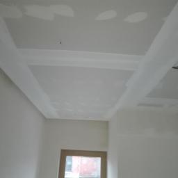 SUFITY - MP MARIUSZ PACZKOWSKI - Sufit w trakcie prac wykończeniowych: widoczne łączenia płyt gipsowo-kartonowych wzmocnione taśmą i pokryte warstwą masy szpachlowej, przygotowane do dalszej obróbki. W tle fragment ściany i okno.