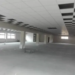 Rozległe, surowe wnętrze biurowe z częściowo zamontowanym sufitem podwieszanym z płyt kartonowo-gipsowych, odsłaniającym instalacje, wsparte na kolumnach, z widocznymi oknami i materiałami...