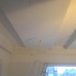 SUFITY - MP MARIUSZ PACZKOWSKI - Wykończone wnętrze z widocznymi elementami dekoracyjnymi na suficie, świeżo pomalowane ściany i sufit w jasnym odcieniu, drabina w tle sugeruje trwające prace wykończeniowe.