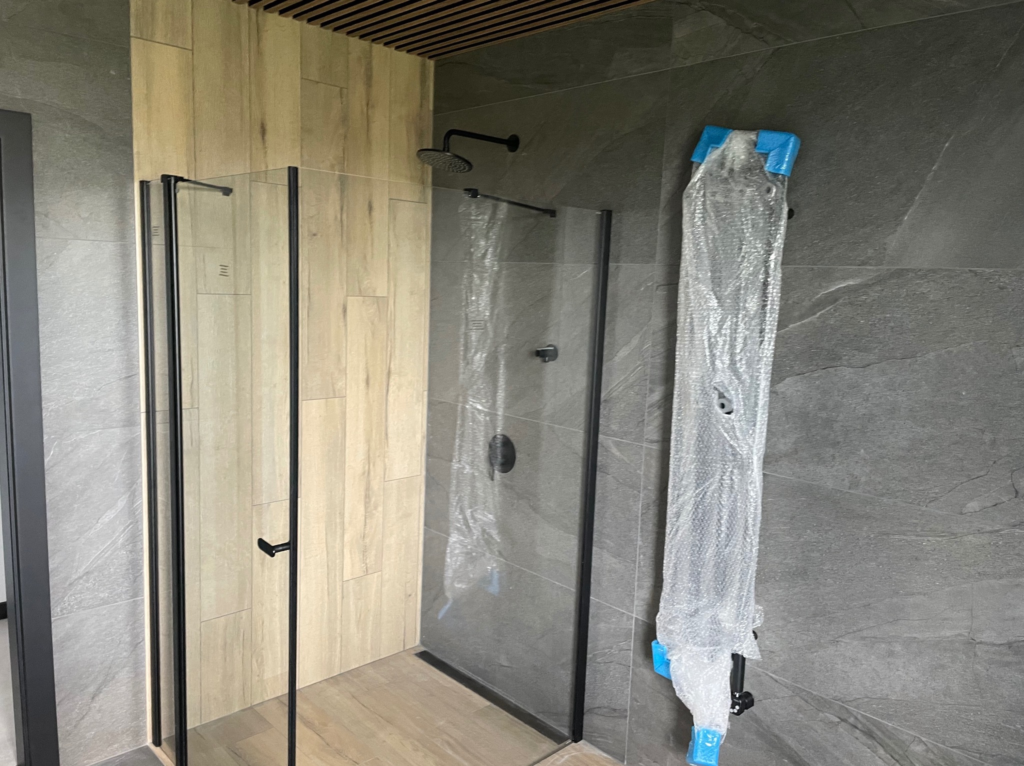 Nowa kabina prysznicowa z czarnymi profilami, częściowo w folii ochronnej, na tle szarych płytek imitujących kamień i drewnopodobnych paneli.