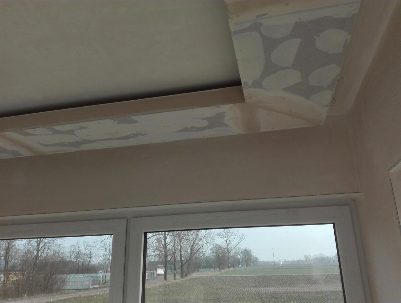 Sufit z widocznymi łatami po szpachlowaniu, przygotowany do dalszych prac wykończeniowych, okno z widokiem na pole i drzewa w tle.